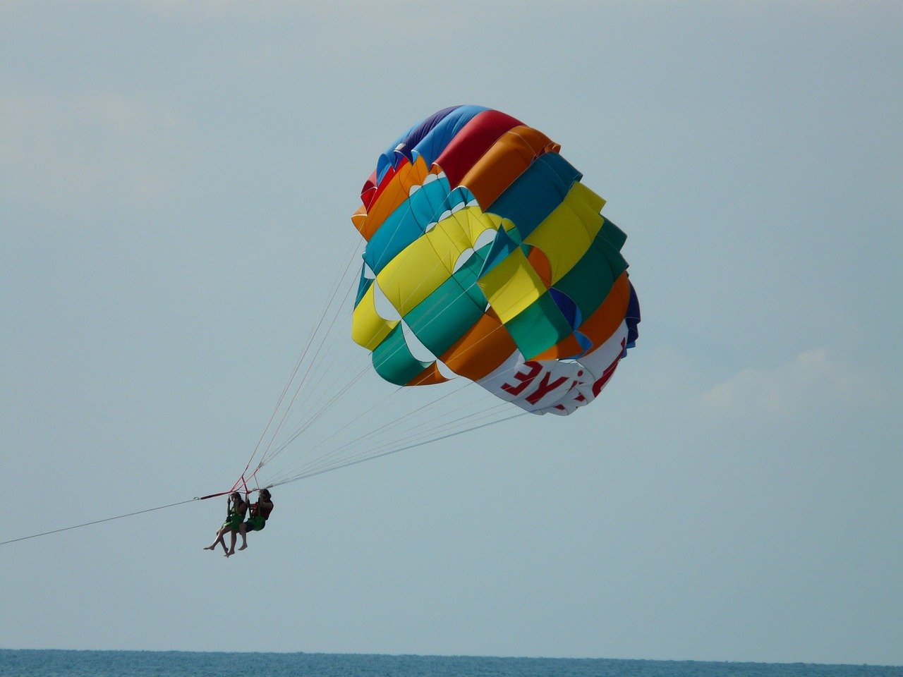 A couple sails on a rainbow parachute against pale blue sky